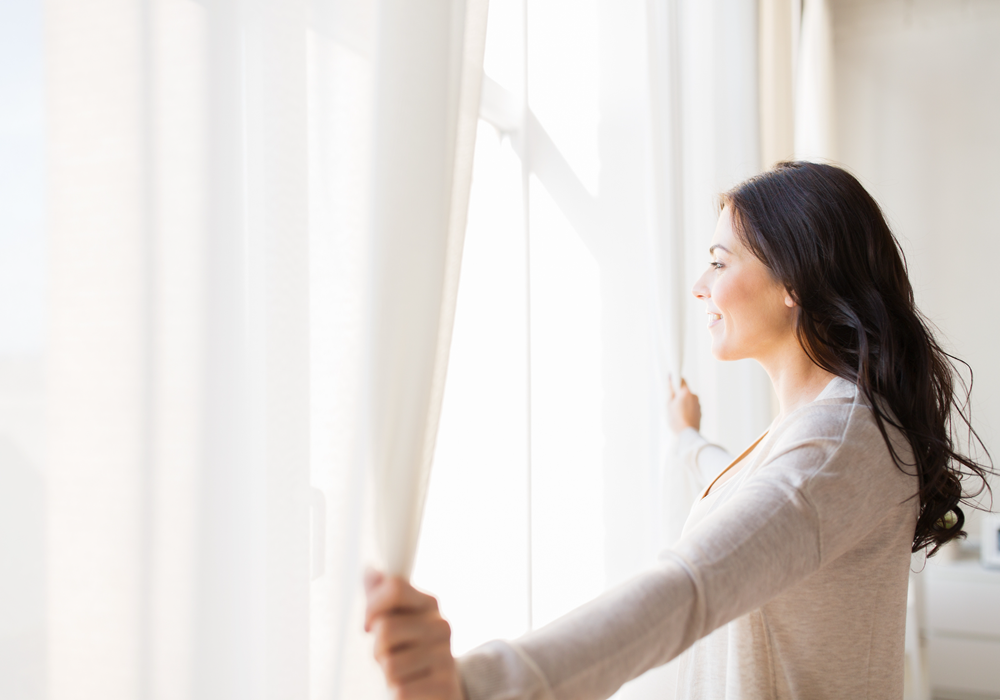 Woman opening up white curtains to let light into apartment home.