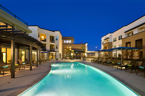Outdoor swimming pool area with sundeck, lounge seating, pergola, and umbrellas at night.